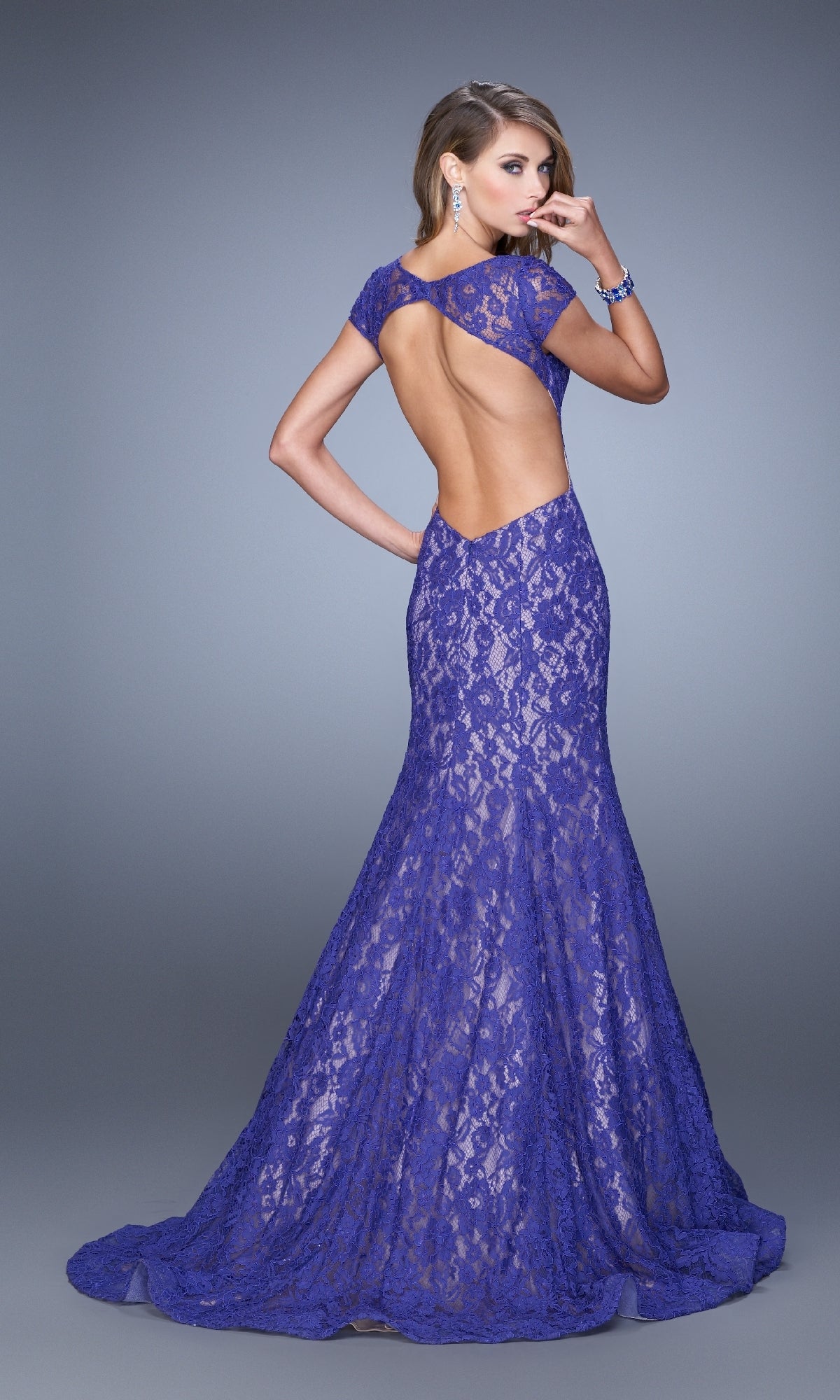 A woman models the Cap-Sleeve Long Lace Prom Dress La Femme 21509, a floor-length royal blue fitted gown with an open back and mermaid skirt, standing in a studio and facing away from the camera.